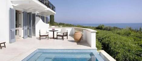Un espace aménagé au bord de la piscine, avec vue sur la mer, un bâtiment blanc doté de volets et des meubles d’extérieur.
