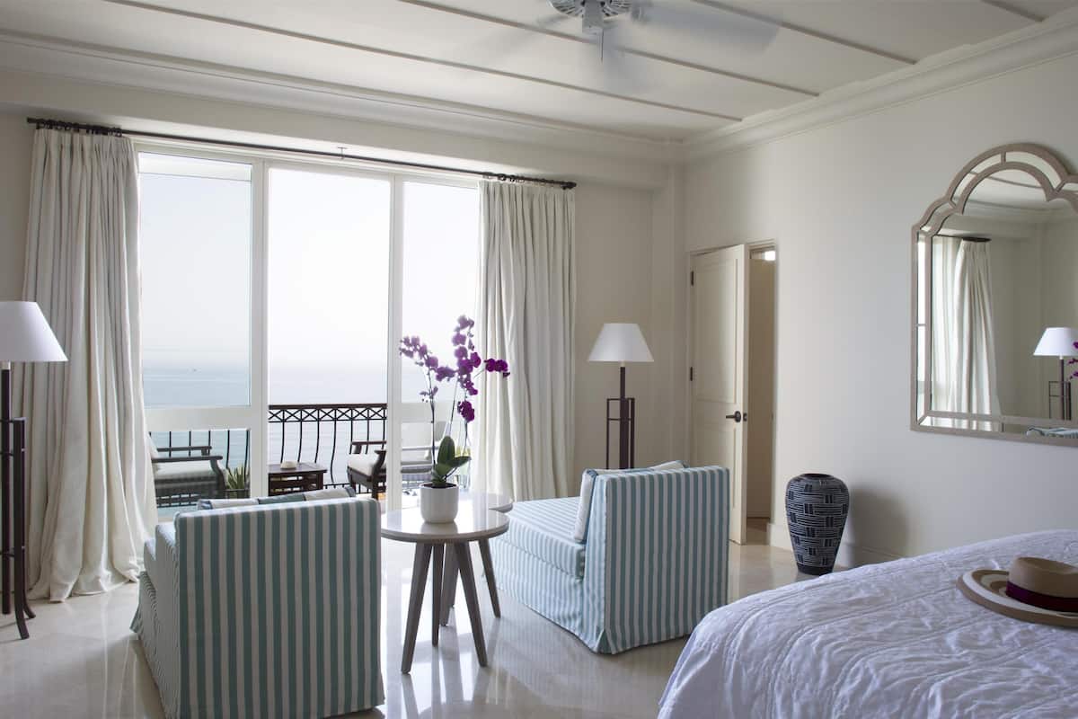 A hotel room with a balcony, a bed, a seating area with a striped sofa and armchairs, a small table with a potted plant, and a large mirror on the wall.