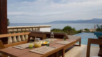 Un espacio de comedor al aire libre con vistas al mar y a las montañas, una piscina y mobiliario de madera.