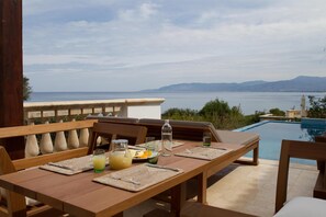 An outdoor dining area with a view of the sea and mountains, a pool, and wooden furniture.