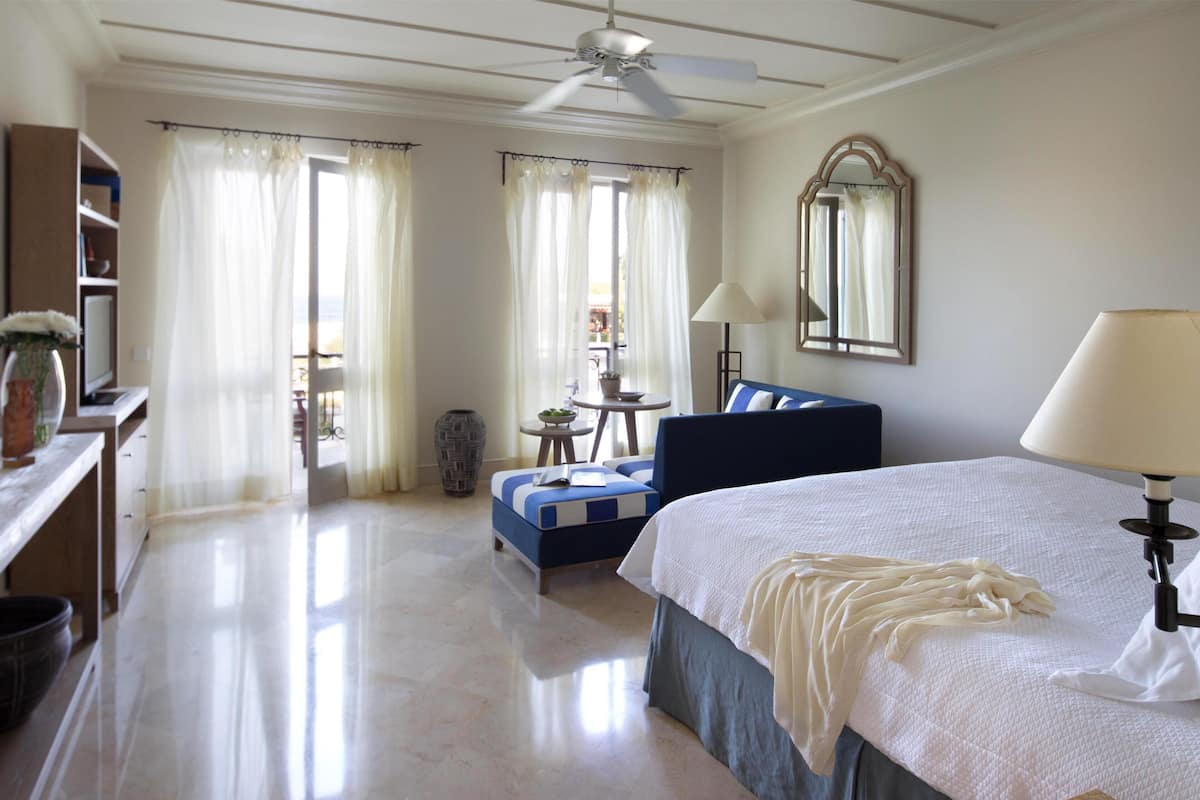 A hotel room with a bed, a desk, a chair, a sofa, a ceiling fan, and a window with curtains.