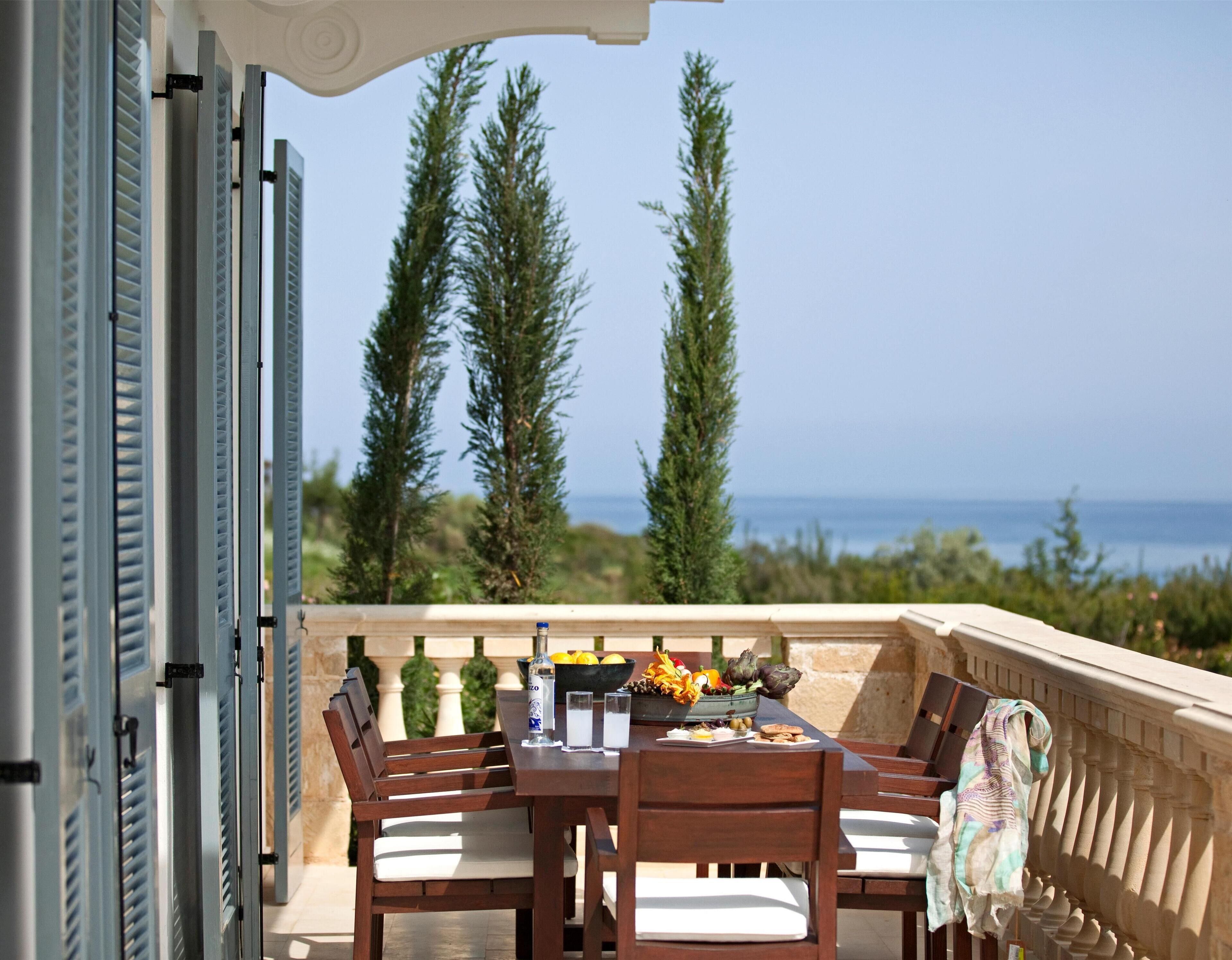 Un espace de restauration en plein air avec une table dressée pour quatre personnes, offrant une vue panoramique sur la mer et les grands arbres.