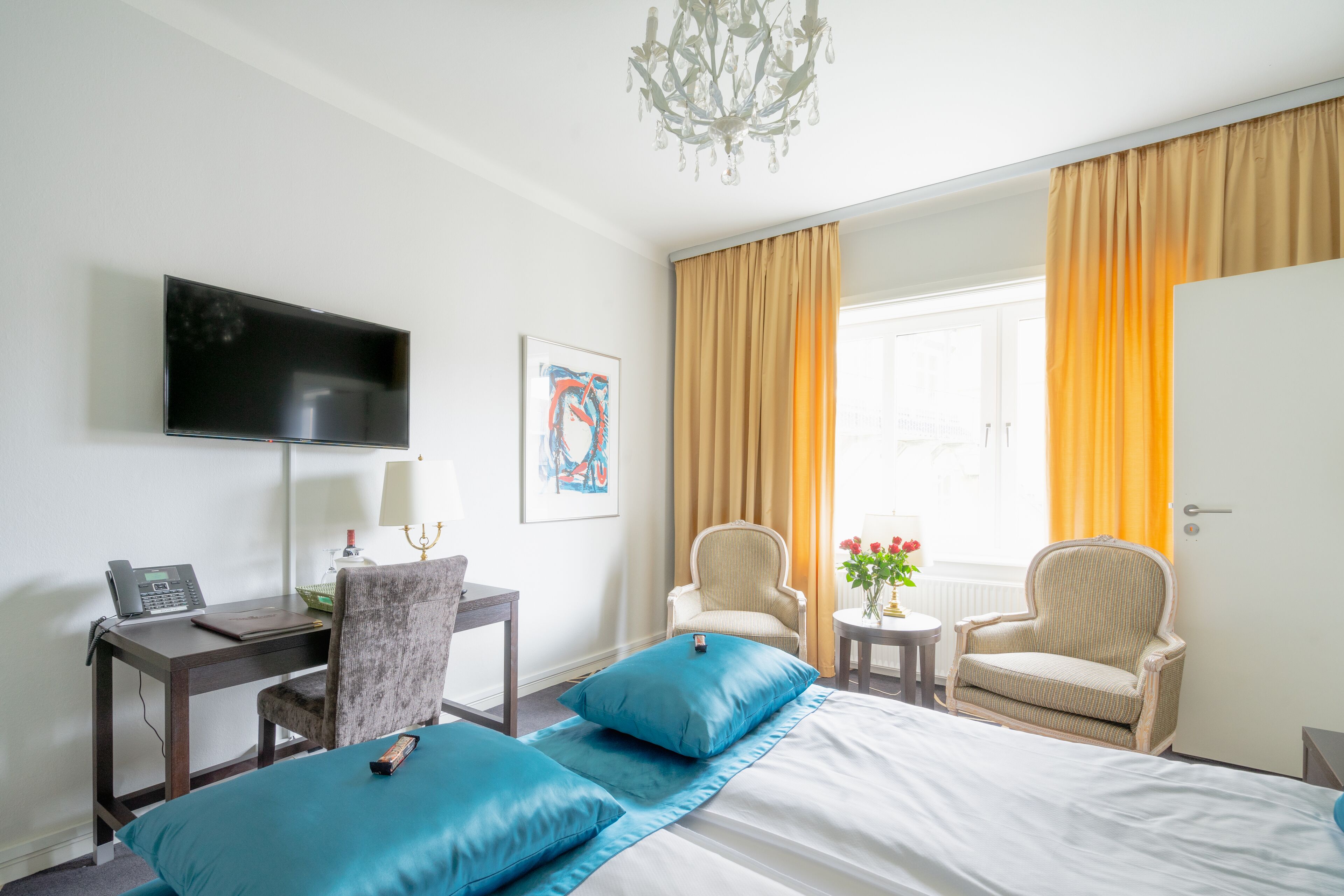 A hotel room with a bed, two armchairs, a desk with a laptop, a TV, and a window with curtains.