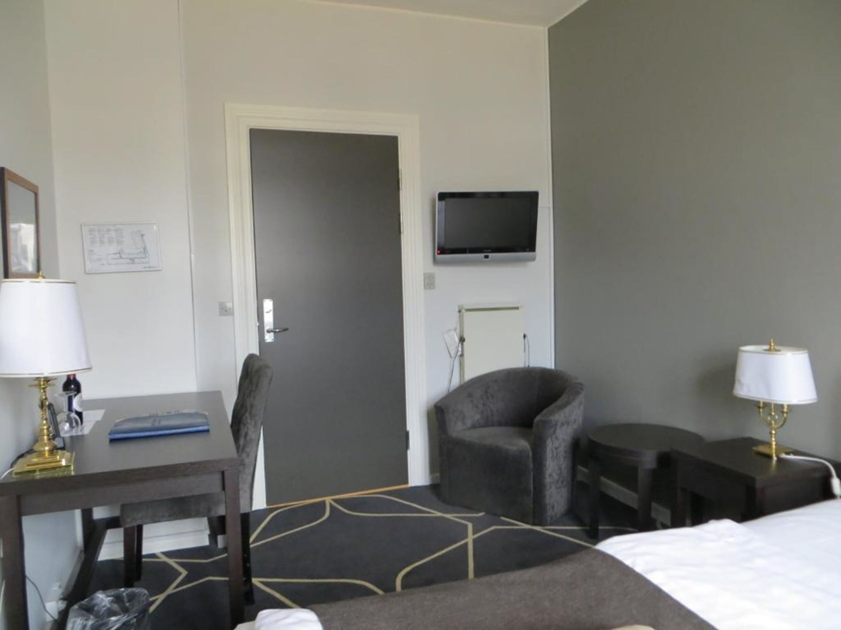 Een hotelkamer met een bed, een bureau met lamp, een stoel, een aan de muur bevestigde televisie en een bijzettafeltje met lamp.