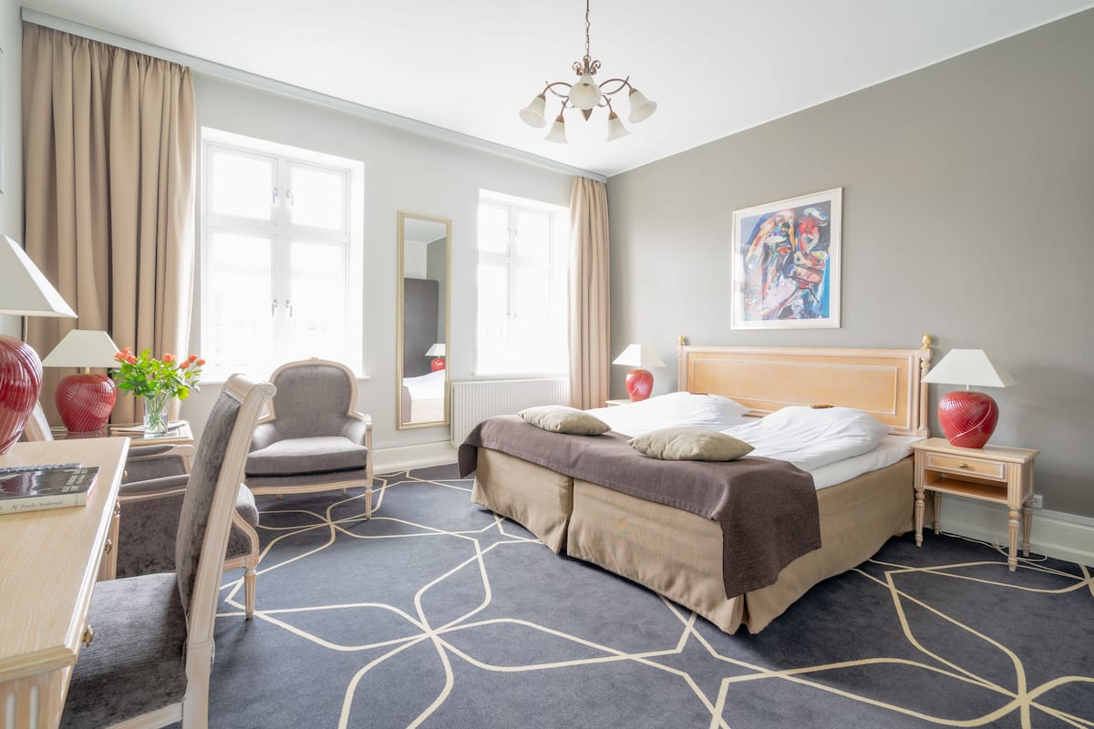 A hotel room with a bed, bedside tables, a desk, a chair, and a large window with curtains.