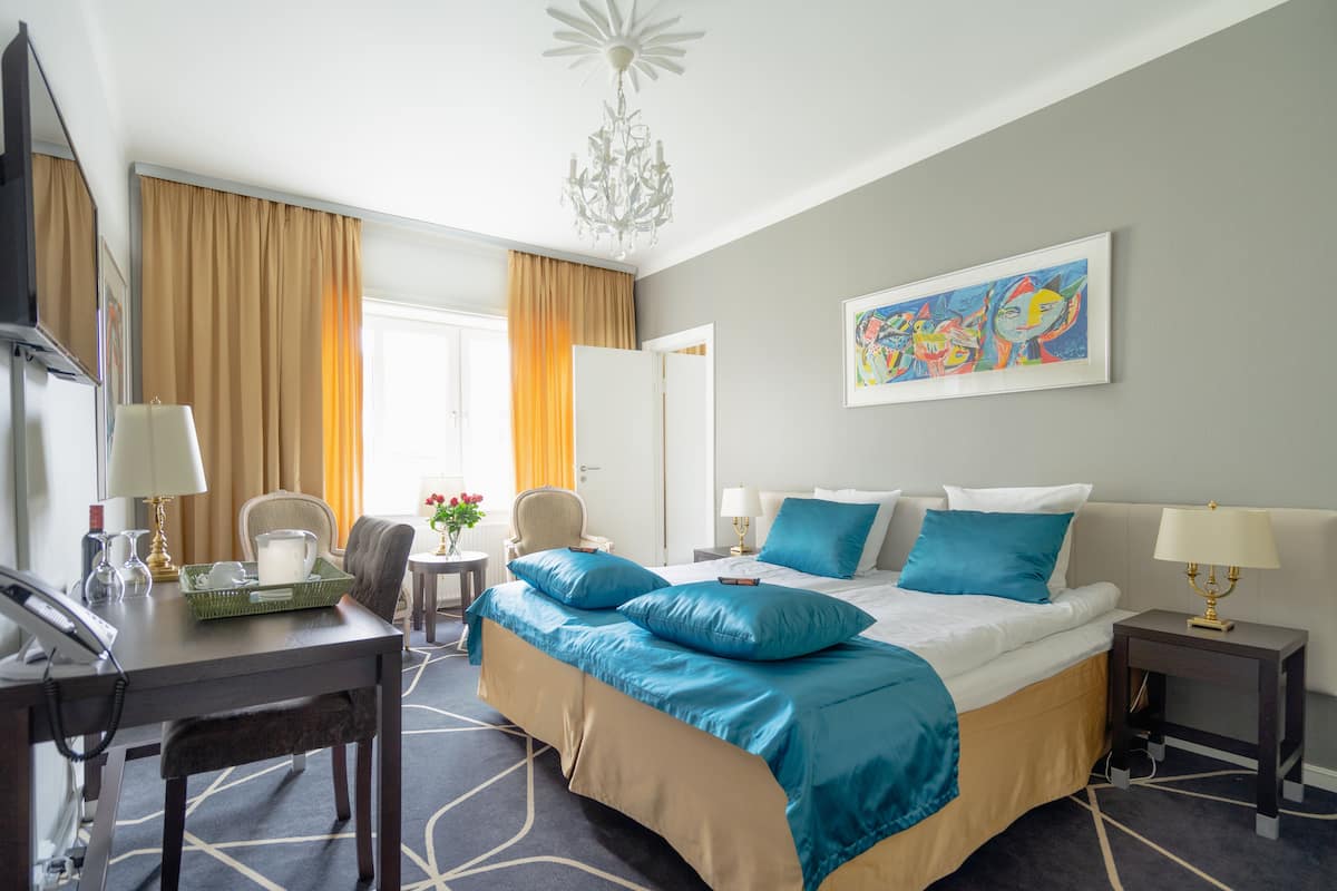 A hotel room with a bed, desk, chair, and a painting on the wall.