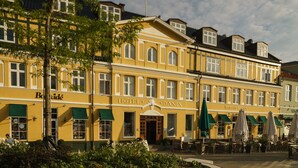 Un bâtiment jaune sur lequel on peut lire les inscriptions « HÔTEL DANIA » et « Jensen’s Badhus » sur le côté. Il y a des tables et des chaises à l’extérieur, et une marquise verte au-dessus de l’entrée.