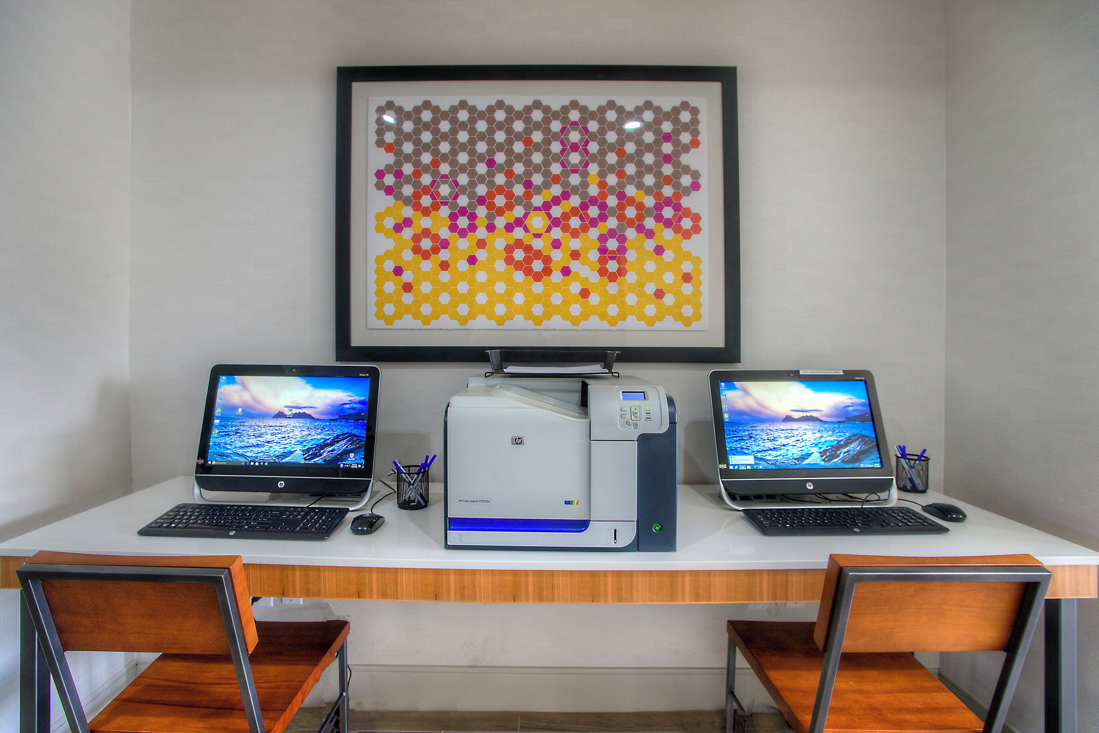 A workspace with two laptops, a printer, and a framed abstract art piece on the wall.