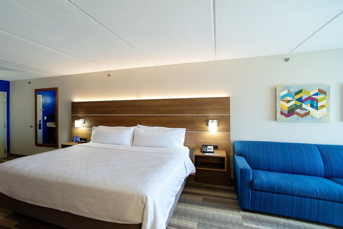 A hotel room with a large bed, bedside tables, a blue armchair, a wooden headboard, a mirror, and a wall art piece.