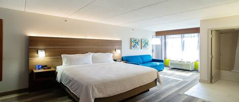 Una habitación de hotel con una cama grande, un sillón azul, una mesita pequeña y una ventana con cortinas.
