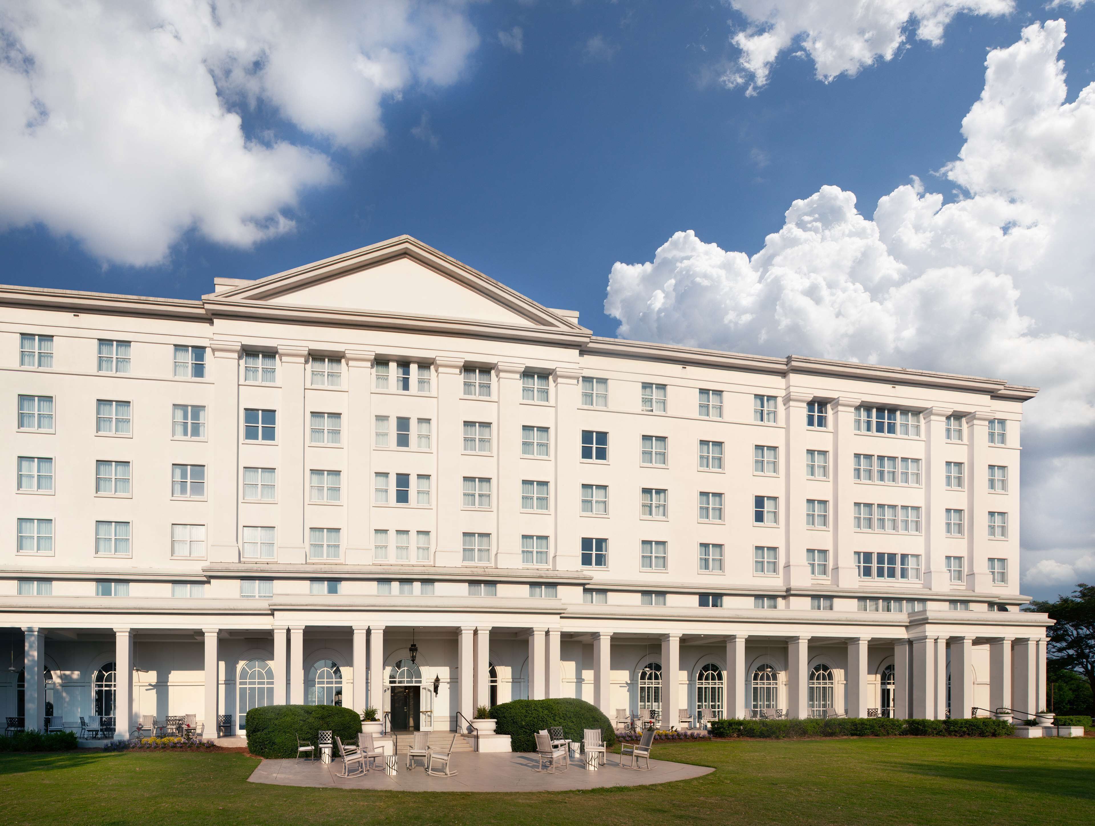 A large, multi-story building with a symmetrical facade, featuring a central entrance with columns, large windows, and a well-maintained lawn with outdoor seating.