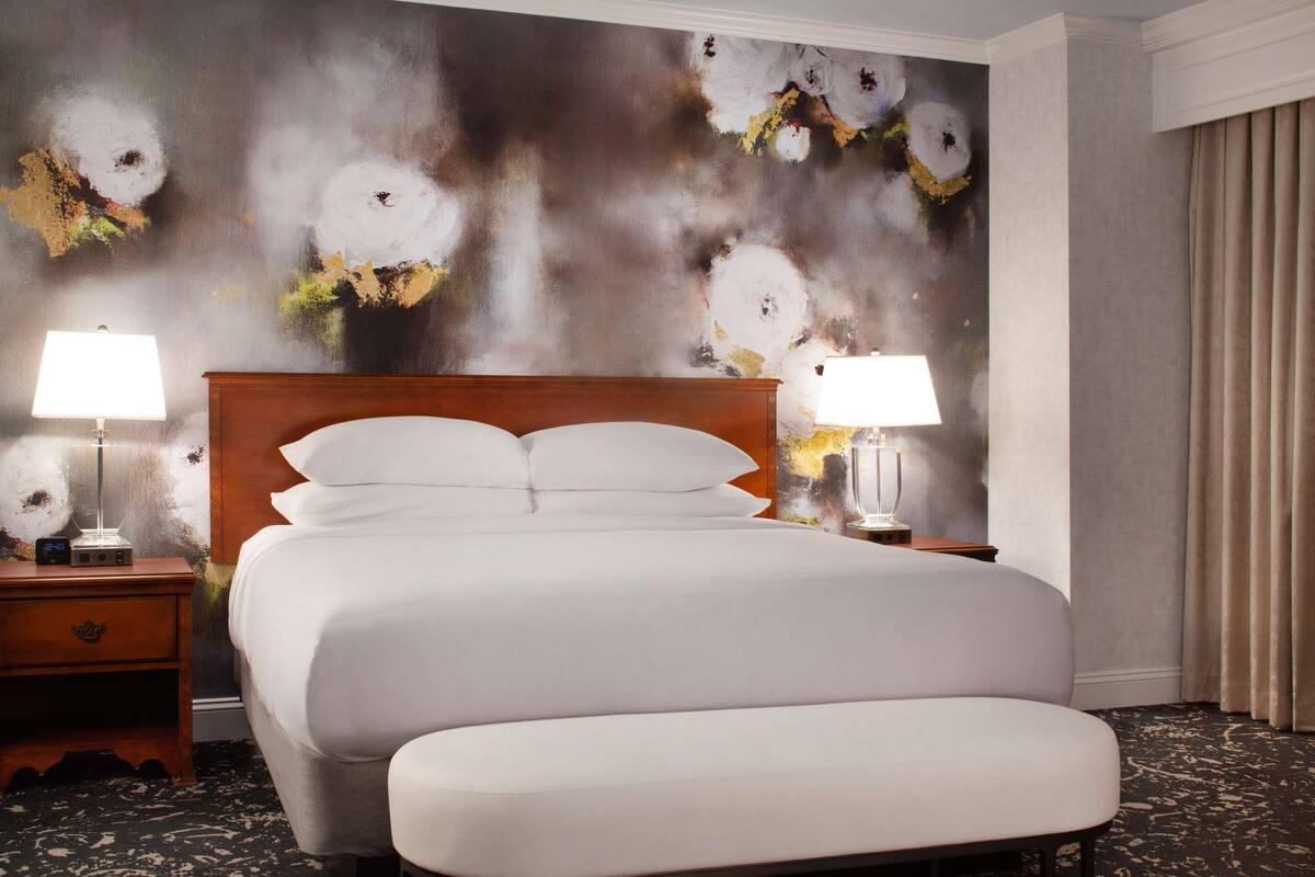 A hotel room with a large bed, two bedside lamps, a wooden headboard, a bench, and a textured wall mural.