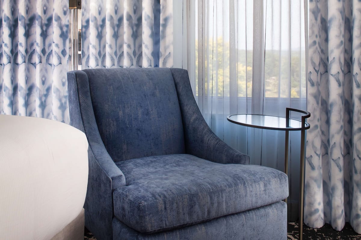 A hotel room with a large bed, a blue armchair, a small round table, and sheer curtains with a leaf pattern.