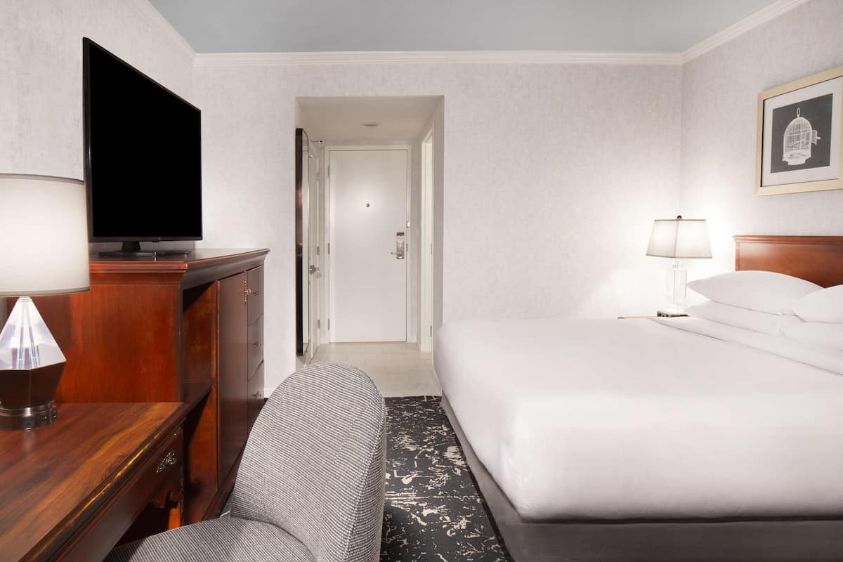 A hotel room with a bed, a desk with a lamp, a television, and a door leading to another room.