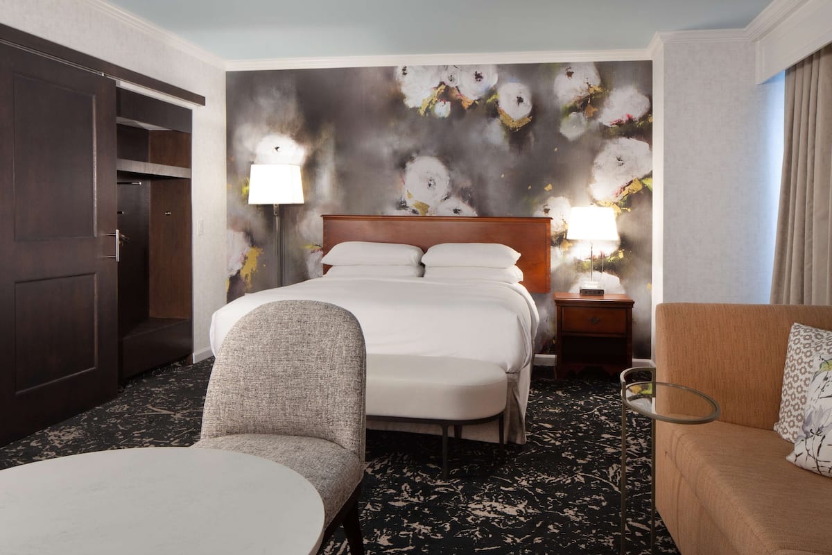 A hotel room with a large bed, a chair, a sofa, a nightstand, and a wall with a floral painting.