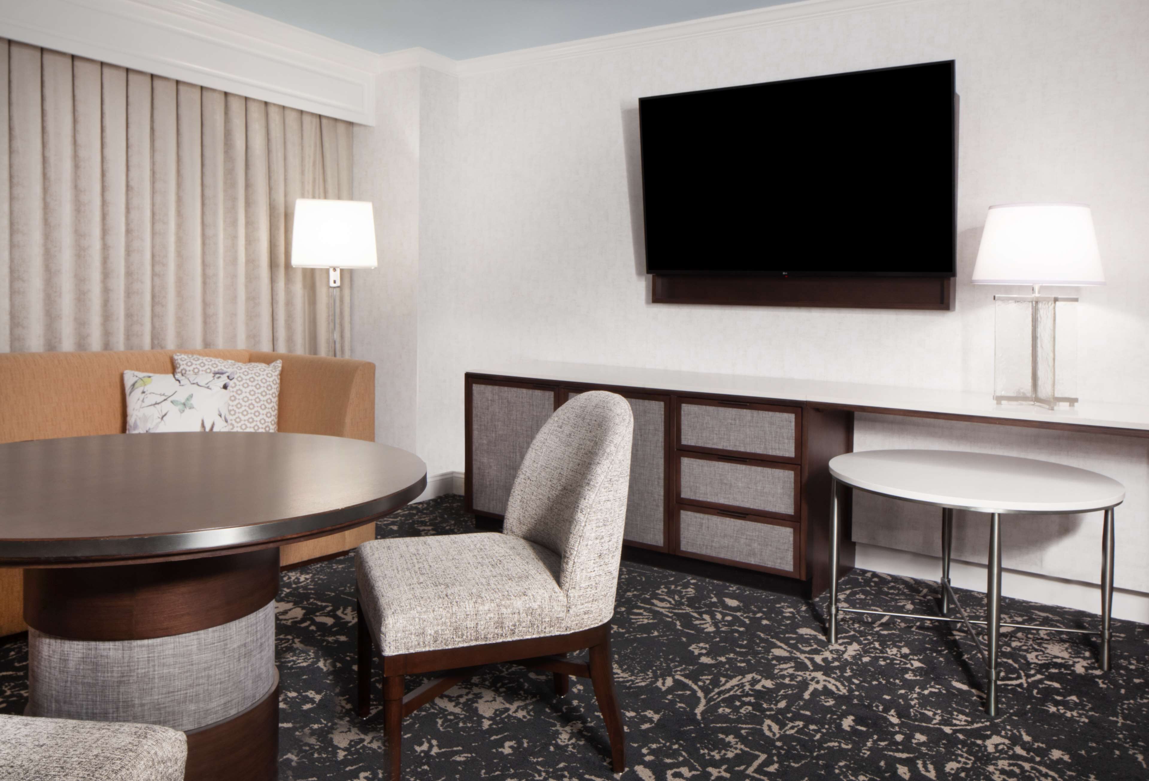 Une chambre d’hôtel comprenant une table à manger ronde, une chaise rembourrée, un canapé avec des coussins à motifs, un téléviseur à écran plat fixé au mur et deux lampes de chevet.
