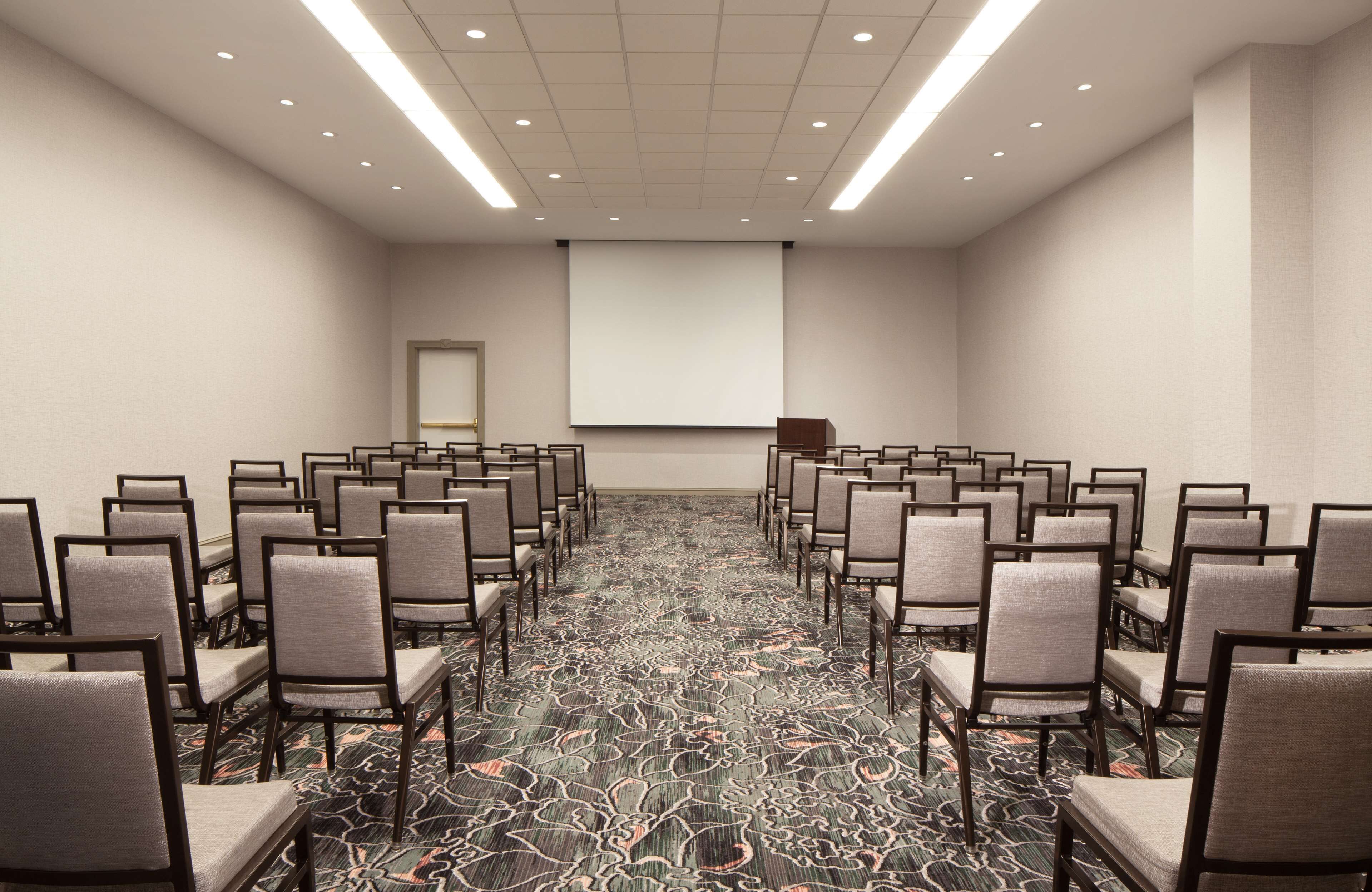 A conference room with rows of chairs facing a projection screen, carpeted floor, and a door on the left.