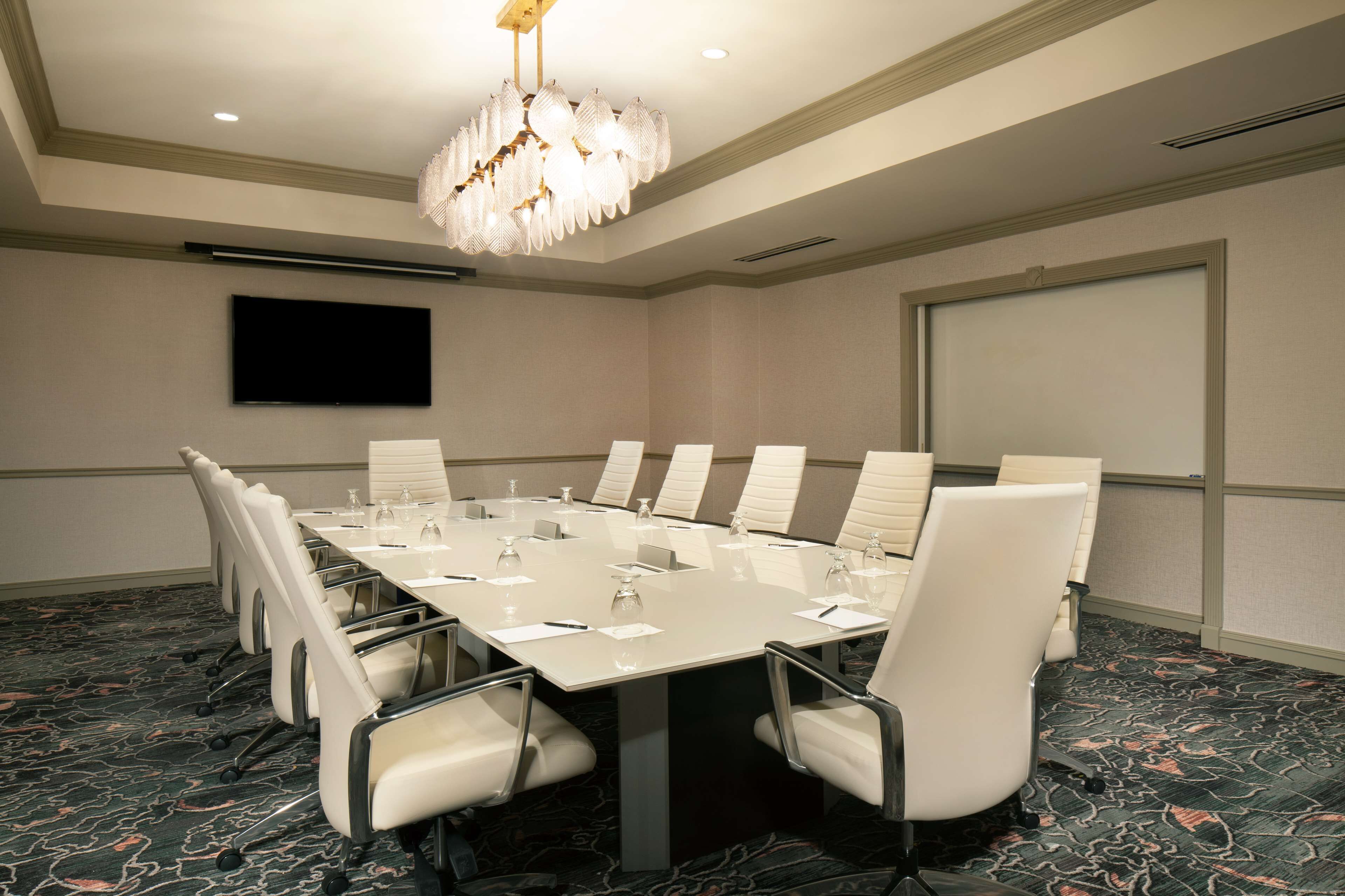 A conference room with a long table, white chairs, a chandelier, a flat-screen TV, and a carpeted floor.