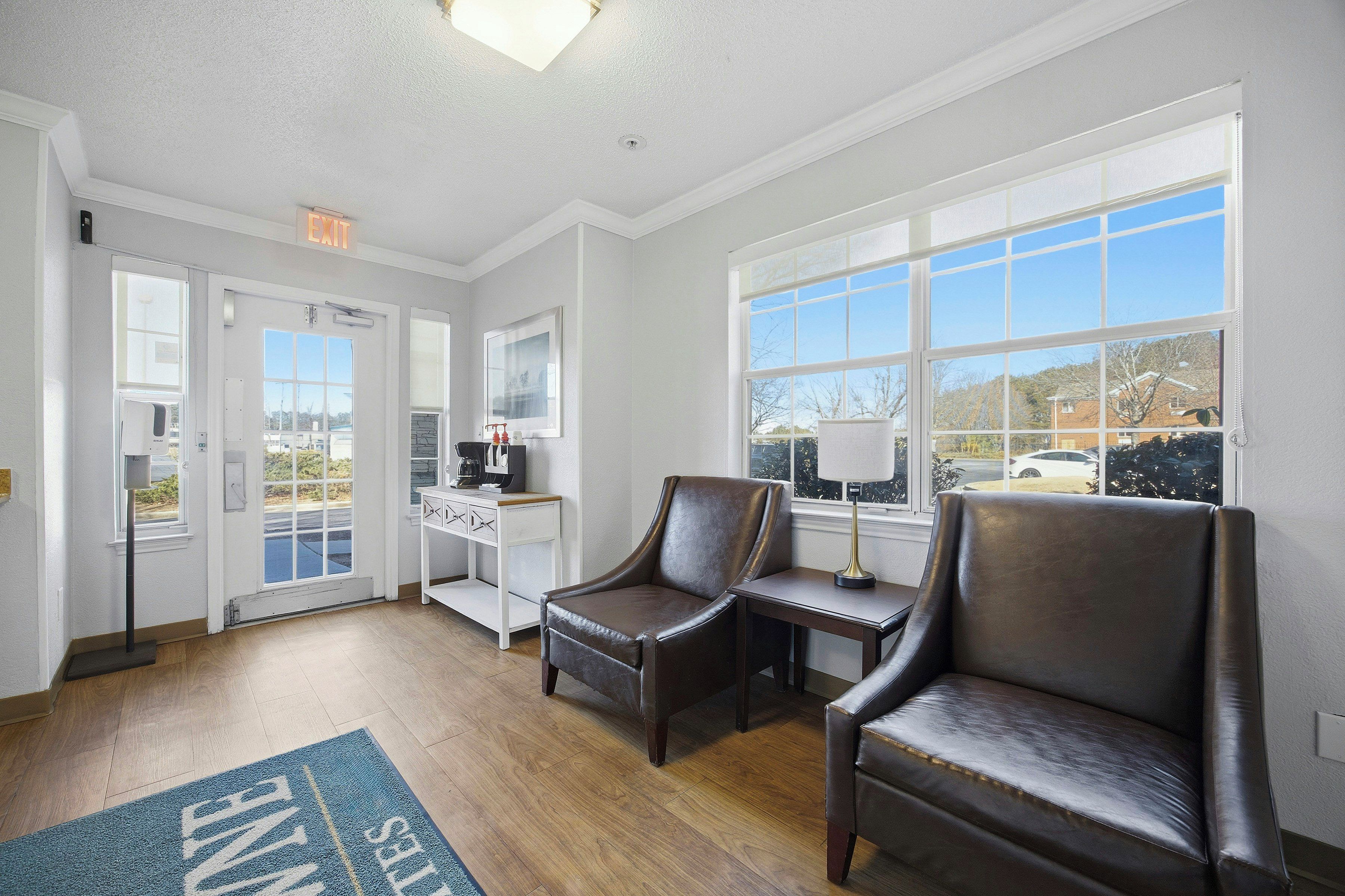 A hotel room with a large window, a desk with a lamp, two leather chairs, and a door leading to a parking area.