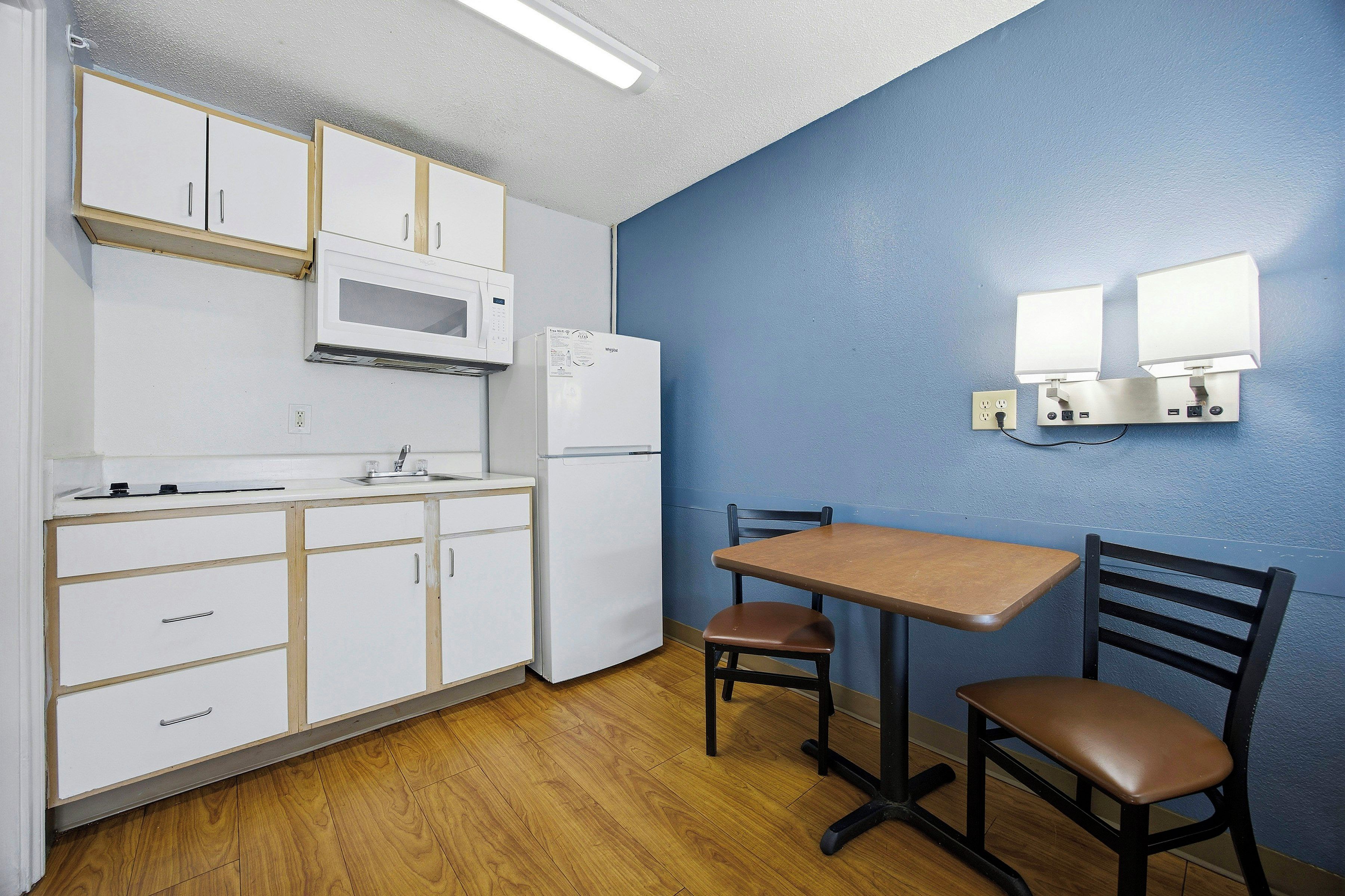 Une petite cuisine équipée avec des meubles blancs, un four à micro-ondes et un réfrigérateur. On y trouve une table avec deux chaises et un luminaire fixé au mur.