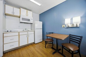 Una kitchenette con armarios blancos, un microondas y un refrigerador. Hay una mesa con dos sillas y una lámpara fijada a la pared.