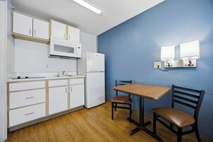 Una kitchenette con armarios blancos, un microondas y un refrigerador. Hay una mesa con dos sillas y una lámpara fijada a la pared.