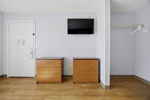 A room with a white door, a flat-screen TV mounted on the wall, two wooden dressers, and a white coat rack. - HomeTowne Studios by Red Roof Atlanta – Peachtree Corners (Peachtree Corners)