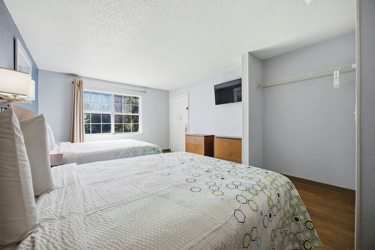 A hotel room with two beds, a window with curtains, a TV mounted on the wall, and a wooden dresser.