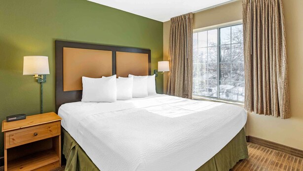 A hotel room with a large bed, two bedside lamps, a nightstand, and a window with curtains. - Extended Stay America Select Suites Chicago Naperville East (Naperville)