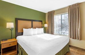 A hotel room with a large bed, two bedside lamps, a nightstand, and a window with curtains. - Extended Stay America Select Suites Chicago Naperville East (Naperville)