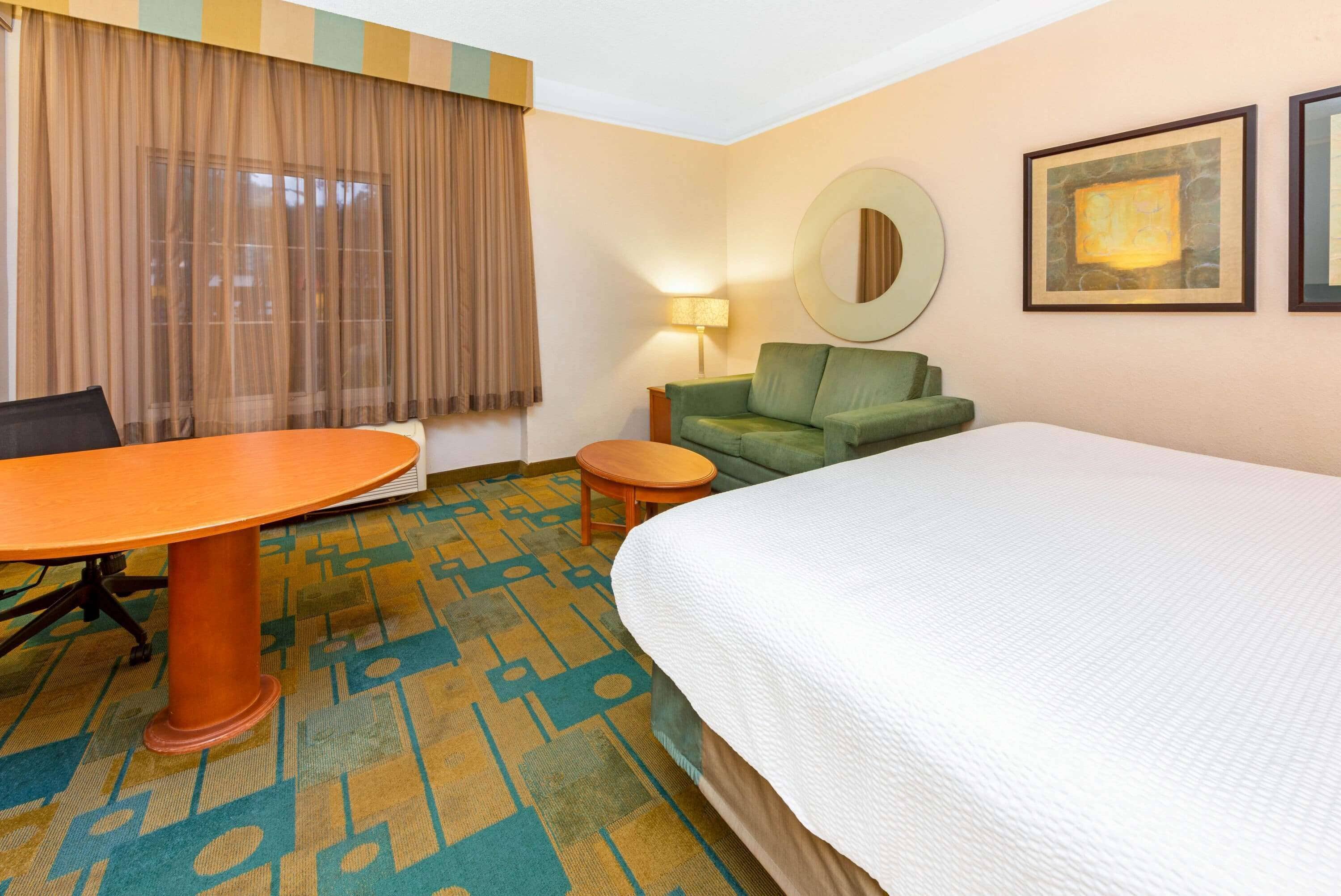 A hotel room with a large bed, a green sofa, a small round table, a desk with a chair, and a patterned carpet.