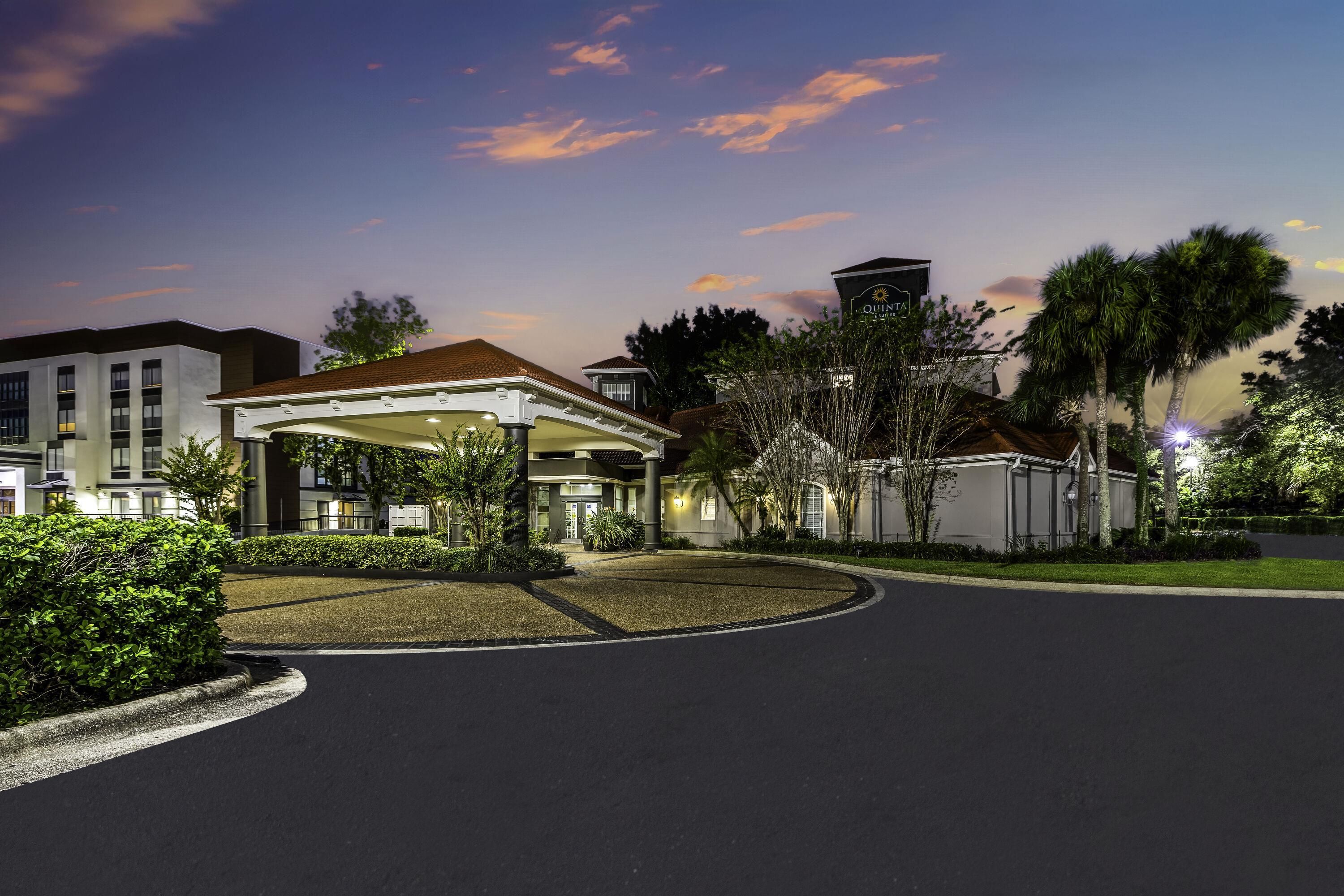 A hotel entrance with a covered walkway, a parking lot, and a landscaped area with trees and shrubs.