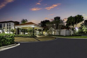 A hotel entrance with a covered walkway, a parking lot, and a landscaped area with trees and shrubs. - La Quinta Inn & Suites by Wyndham USF (Near Busch Gardens) (Tampa)