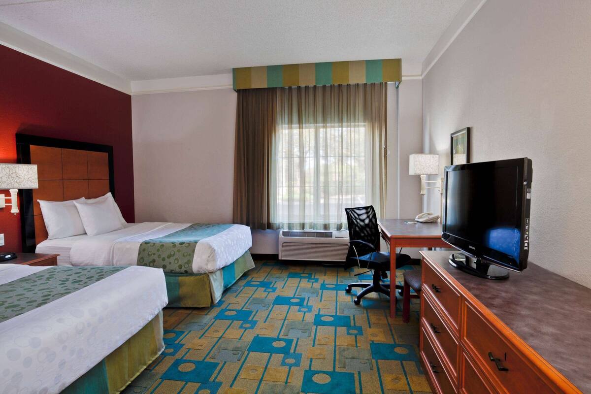 A hotel room with two beds, a television, a desk with a chair, and a window with curtains.