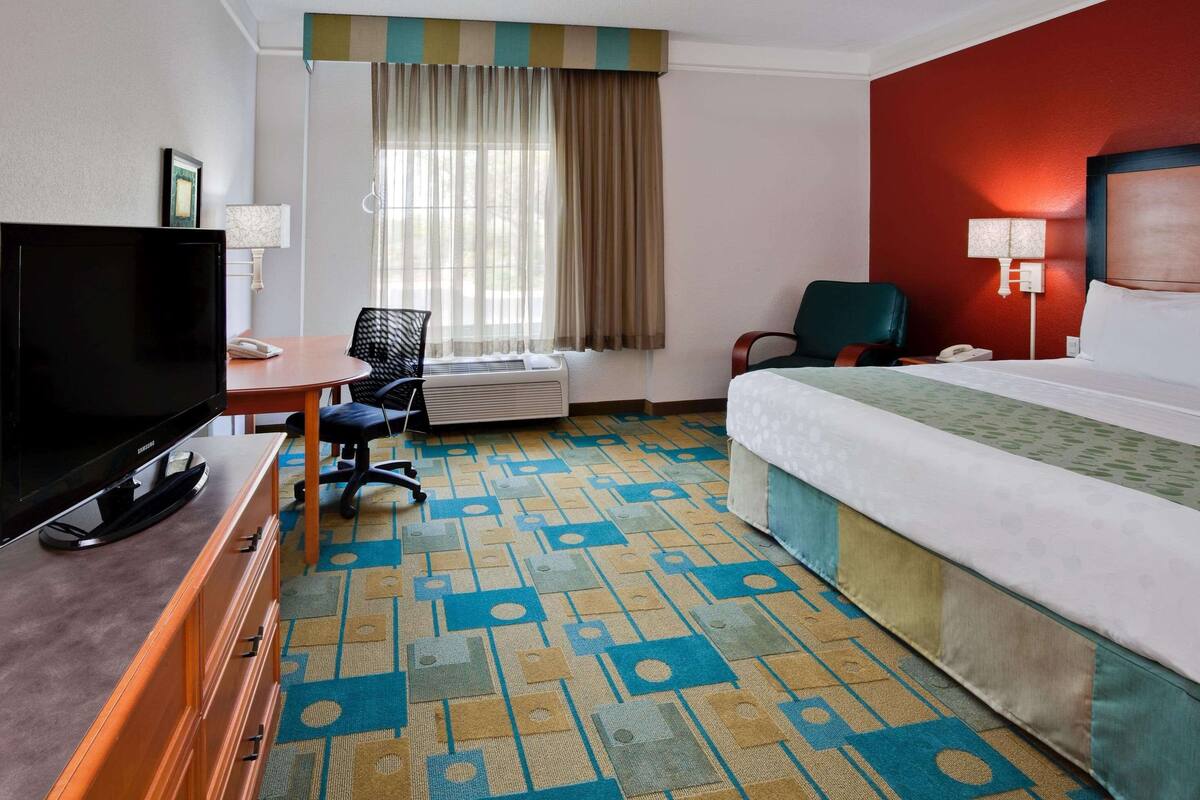 A hotel room with a bed, a desk with a television, a chair, a desk lamp, and a window with curtains.