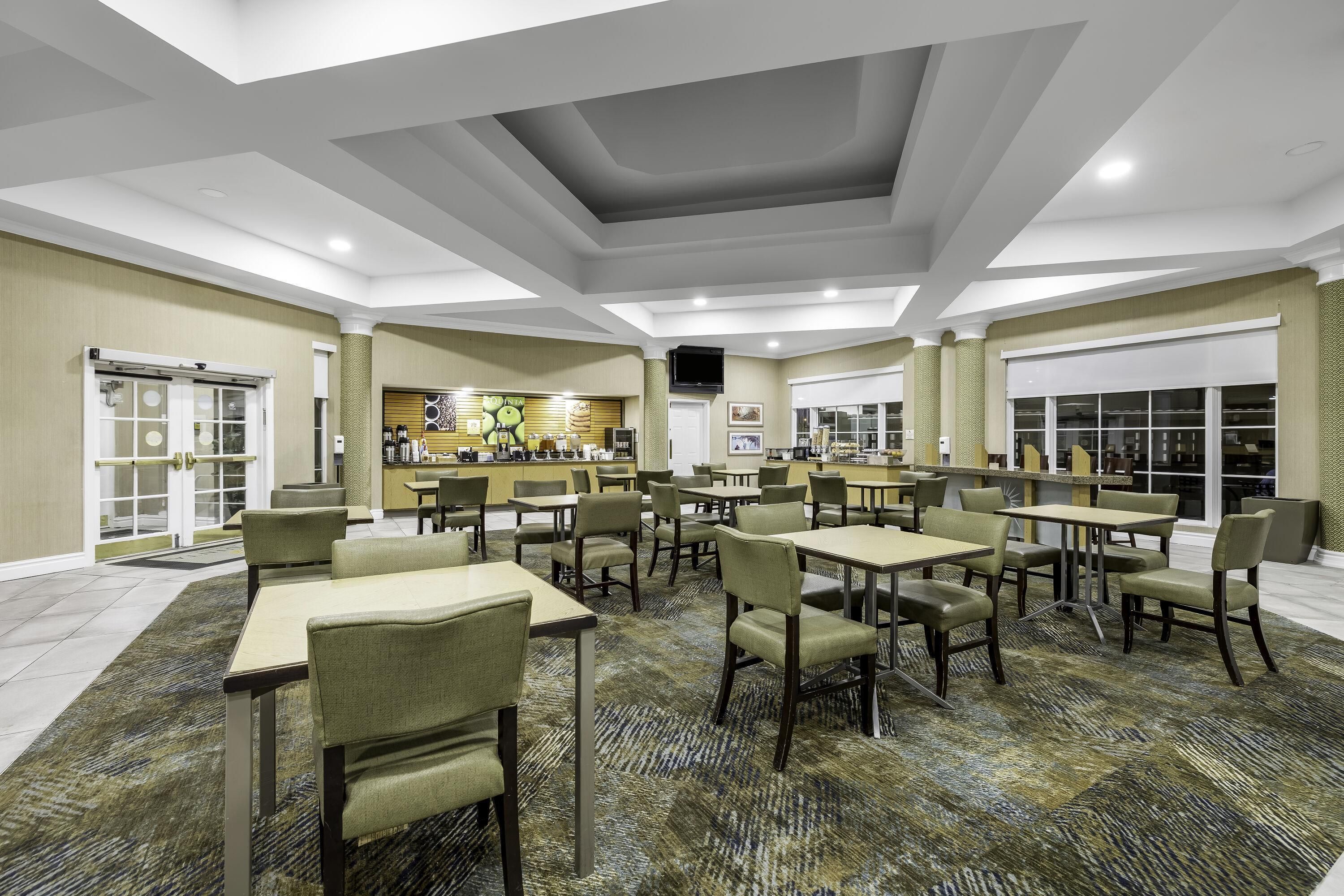 A hotel dining area with multiple tables and chairs, a kitchen in the background, and a patterned carpet.