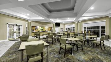 A hotel dining area with multiple tables and chairs, a kitchen in the background, and a patterned carpet.