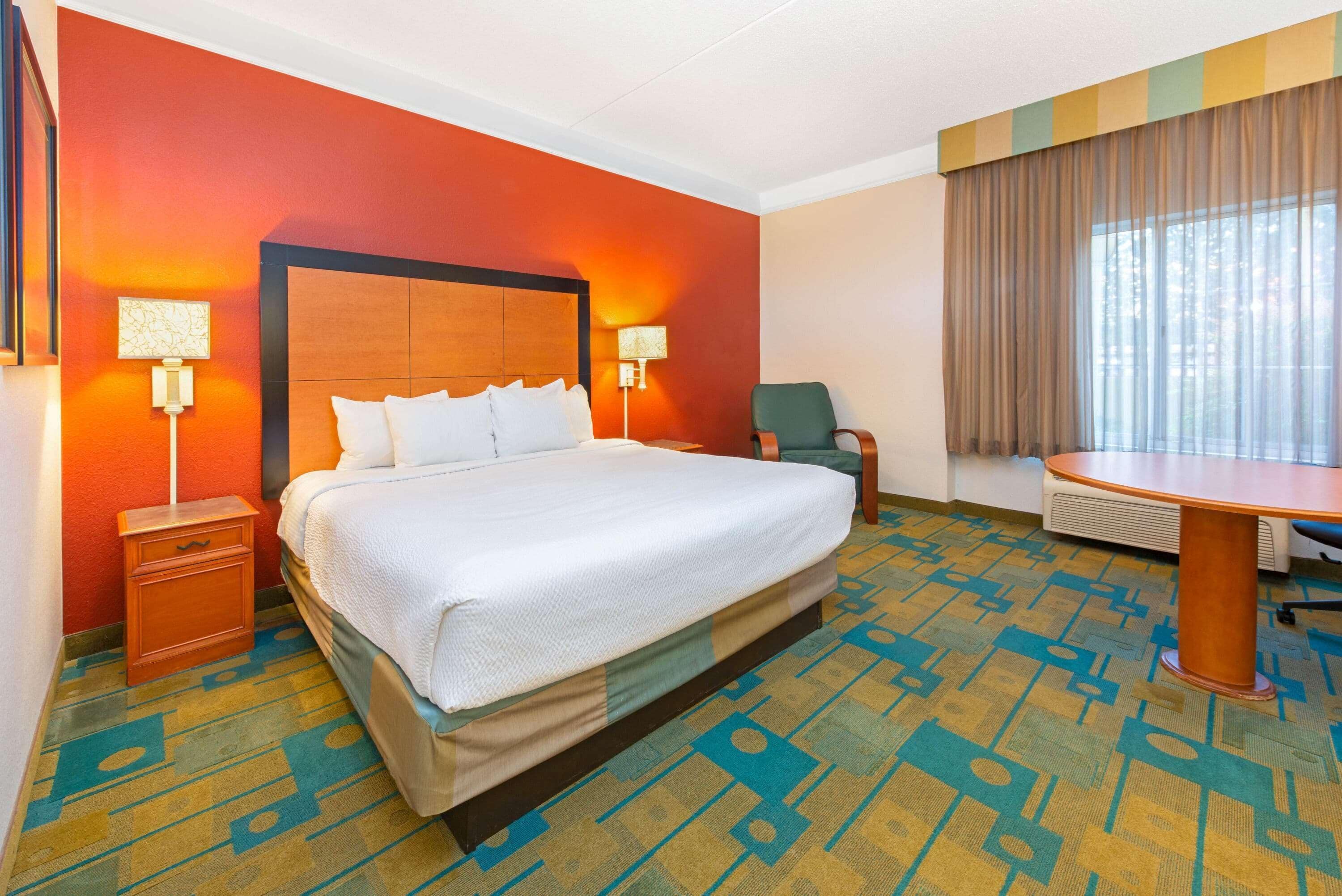 A hotel room with a large bed, two bedside tables with lamps, a chair, a desk, and a large window with curtains.