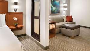 Una habitación de hotel con cama, zona de estar con sofá y reposapiés, una mesita con una planta y un cuadro en la pared.