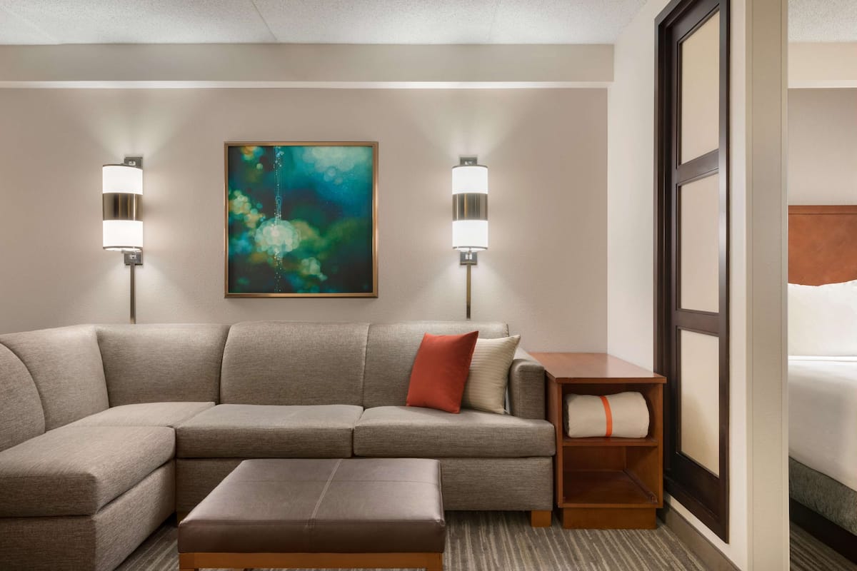 A hotel room with a sectional sofa, a small ottoman, a wooden side table, and a painting on the wall.