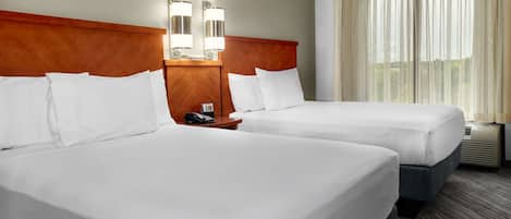 Quarto de hotel com duas camas, uma cabeceira de madeira, uma mesa de cabeceira com telefone e uma janela com cortinas translúcidas.