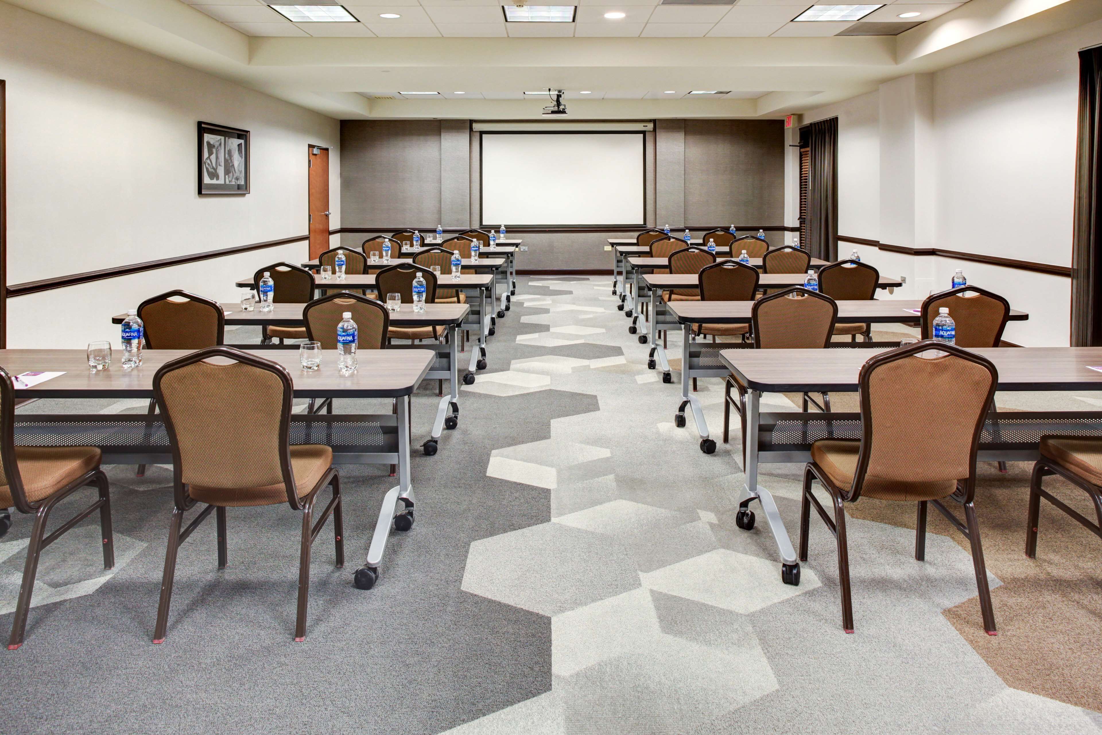 A conference room with rows of tables and chairs, equipped with water bottles, a projector screen, and a presentation screen.
