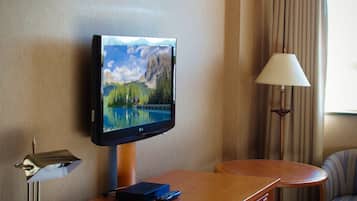 Habitación de hotel con televisor de pantalla plana montado en una cómoda de madera, una lámpara, una silla y una ventana con cortinas.