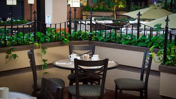 An outdoor dining area with tables and chairs, surrounded by lush greenery and decorative lighting.
