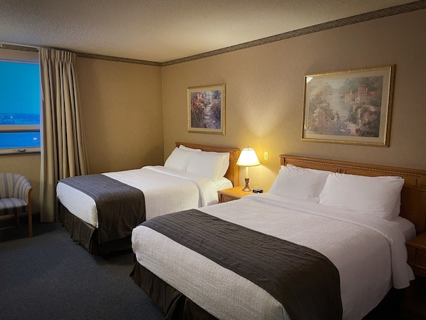 A hotel room with two beds, a chair, a lamp, and two paintings on the wall.
