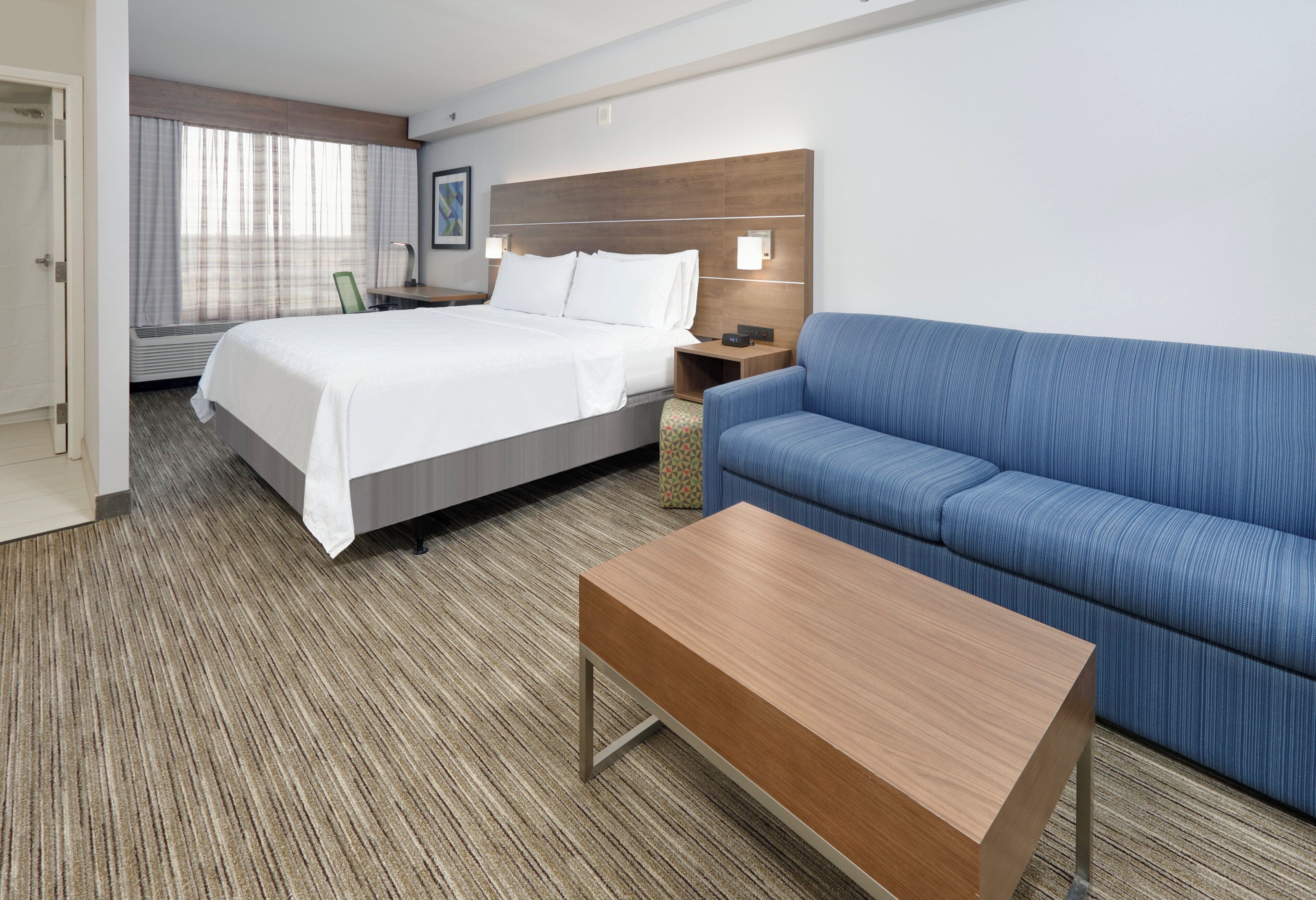 A hotel room with a bed, a blue sofa, a wooden coffee table, a desk, and a wall-mounted lamp.