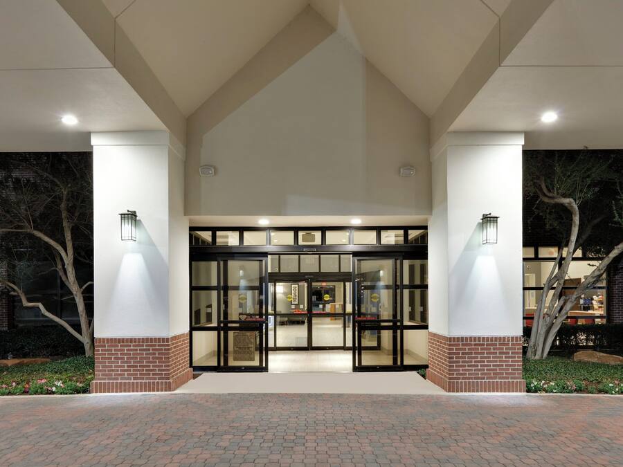 A building entrance with a glass door and brick pillars, surrounded by a paved area and trees.