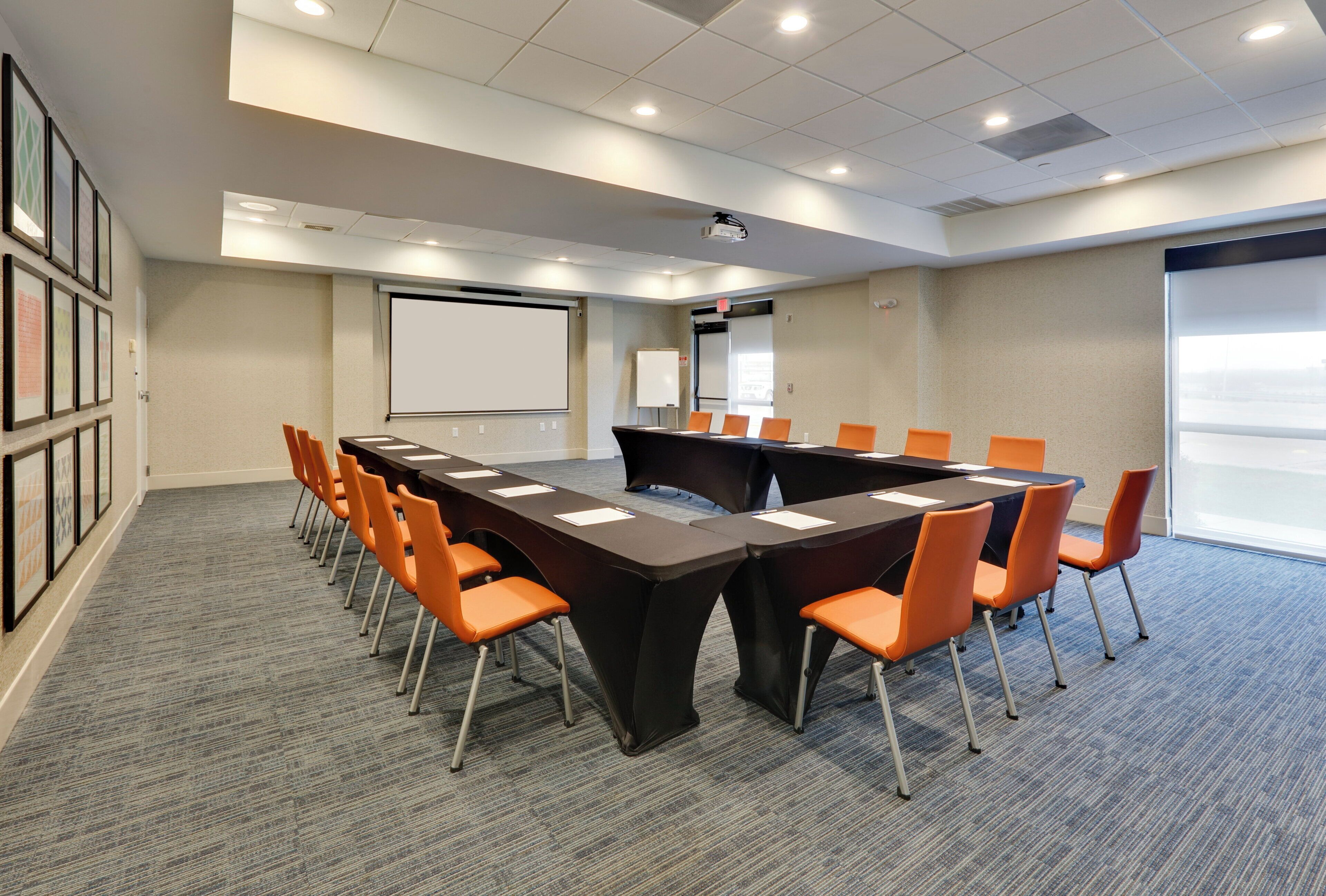 A conference room with a long table, orange chairs, a projector, a whiteboard, and framed pictures on the wall.