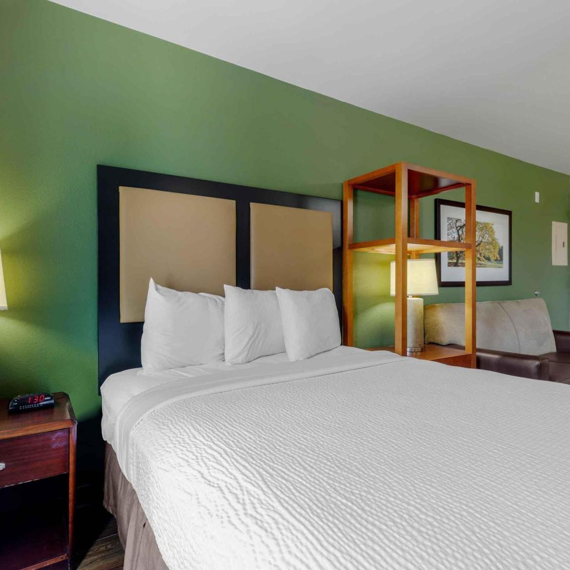 Une chambre d’hôtel avec un grand lit, une tête de lit en bois, une table de chevet avec une horloge et un tableau encadré au mur.