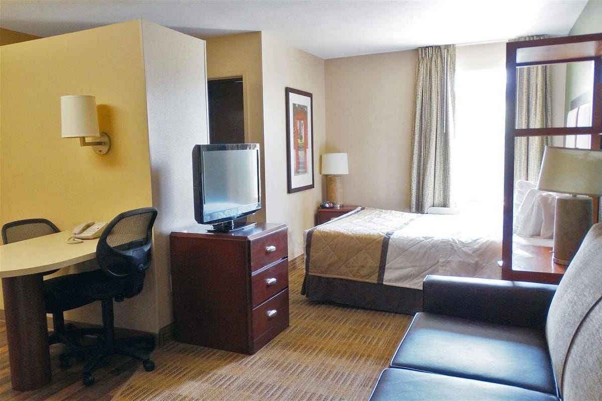 A hotel room with a bed, a desk with a chair, a television, a nightstand, and a window with curtains.